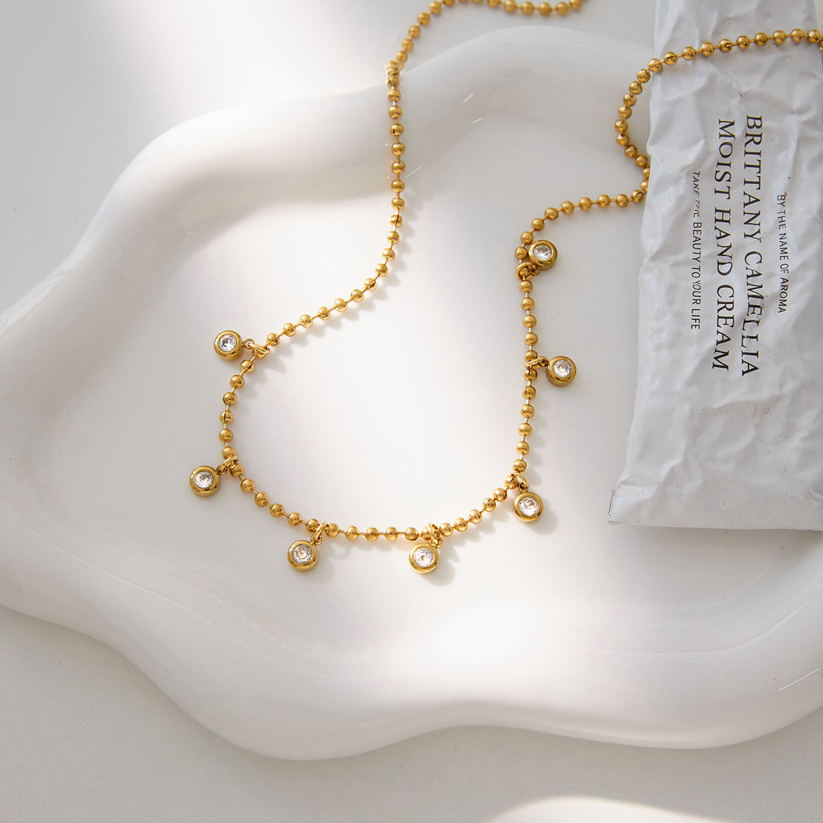 Gold beaded necklace on a white surface with a hand cream packet.