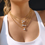 A close-up of a model wearing a multi-layered gold necklace with clear teardrop and oval gemstones, paired with a white tank top.