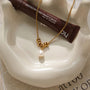 A gold chain necklace with a cluster of gold beads and a single white pearl pendant, displayed on a white ceramic dish next to a brown tube of lip tint.