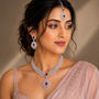 A woman wearing a traditional Indian jewelry set featuring a silver-toned necklace, matching drop earrings, and a forehead piece adorned with deep red gemstones, paired with a light pink embroidered sari.