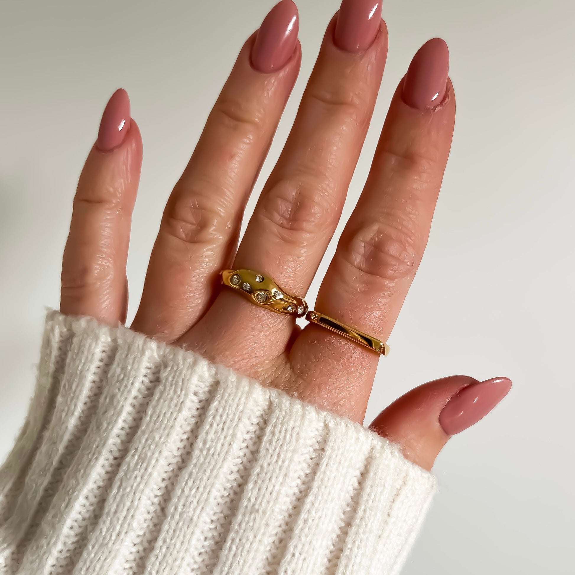 Hand wearing a gold ring with a white sleeve on a neutral background
