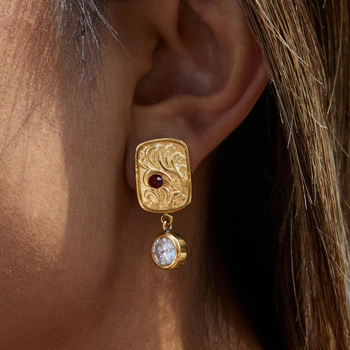 Gold earring with a red gemstone and clear stone worn by a person.